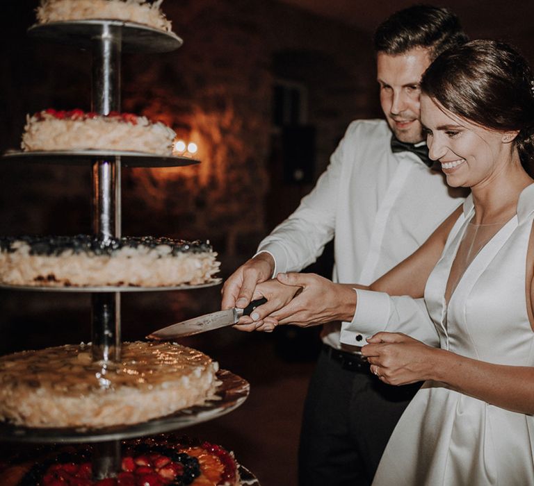 Smiling bride in white Jesus Piero wedding dress cuts five-tiered wedding cake with groom at German wedding