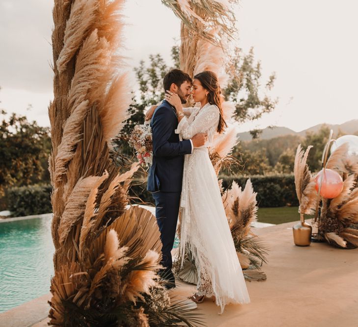 Bride in a lace Marylise Bridal gown with tassels kissing her husband at the poolside ceremony with pampas grass decor 