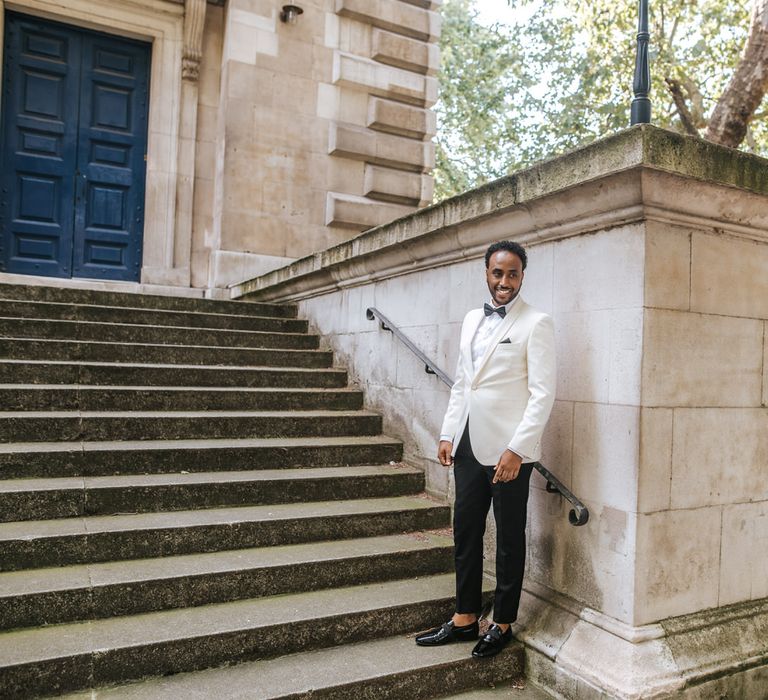 Stylish groom in a white tuxedo jacket and bow tie standing behind a wall waiting for his bride 