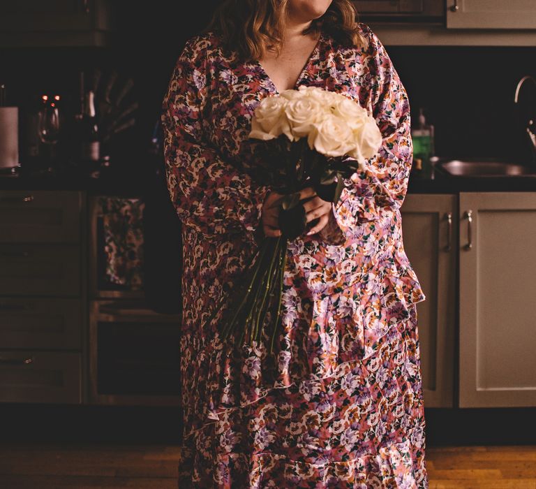 Bride holds long stemmed white rose bouquet whilst she wears floral ASOS wedding gown