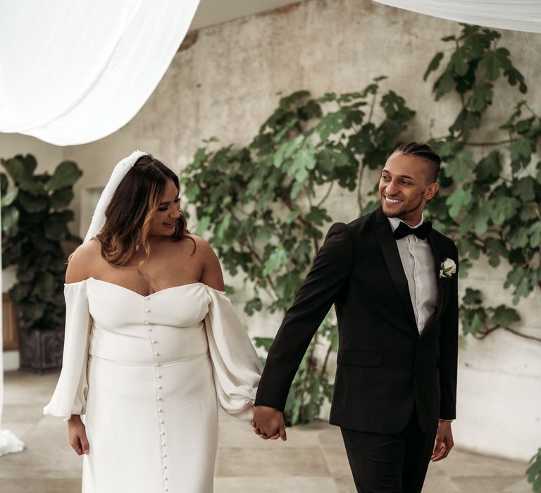 Black tie wedding with the bride in a strapless wedding dress and button detail holding her grooms hand in a tuxedo suit