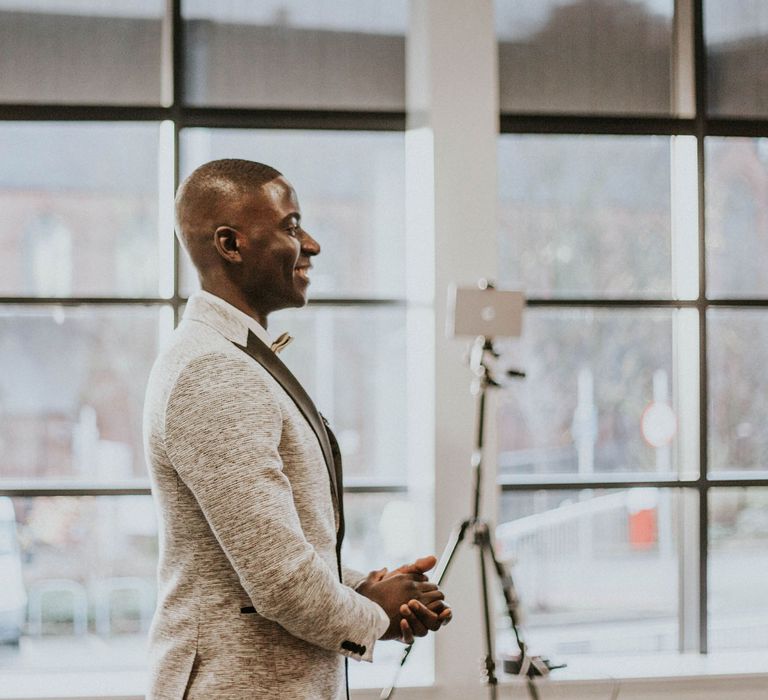 Smiling groom in grey Moss Bros suit with black lapels holds hands together as he sees bride for the first time at Bridge Community Church wedding