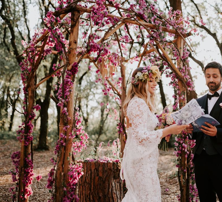 Bride in long sleeved lace boho wedding dress and flower crown reading does with groom in navy suit and bow tie in front of wooden wedding arch decorated with pink flowers