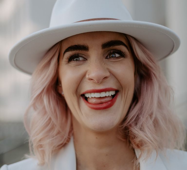 Close up of white bride with pink hair smiling with red lip and nose piercings also wearing a fedora hat 