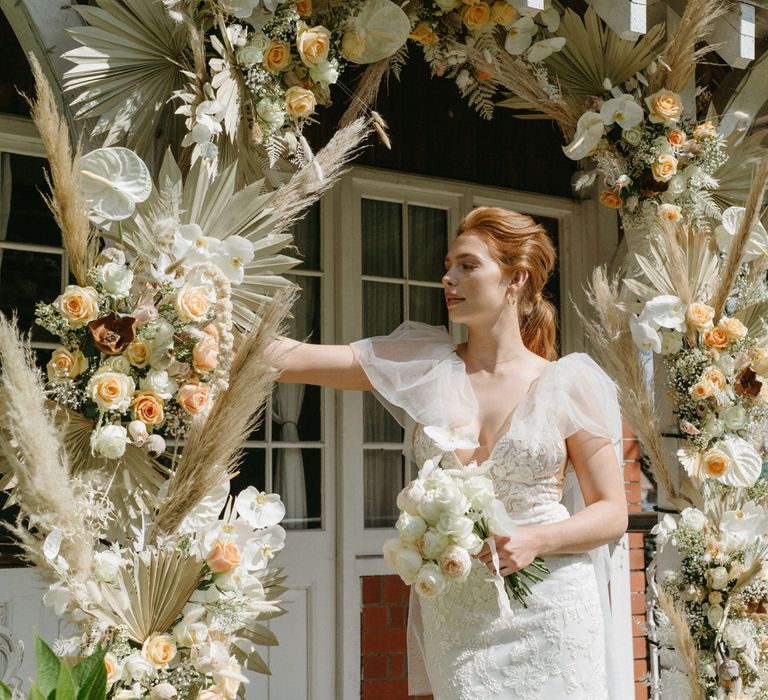 Boho bride at Berwick Lodge with puff cap sleeve wedding dress standing under an arch of dried palms, pampas grass, white anthuriums, roses and orchids