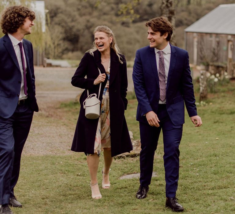 Three laughing wedding guests, two in navy suits and one in midi dress and coat walking towards garden wedding at Hayne, Devon