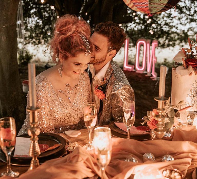 Bride with pink curled hair and hair gems in gold sequinned wedding dress sits at outdoor wedding table with groom in silver sequinned suit jacket at festival themed wedding