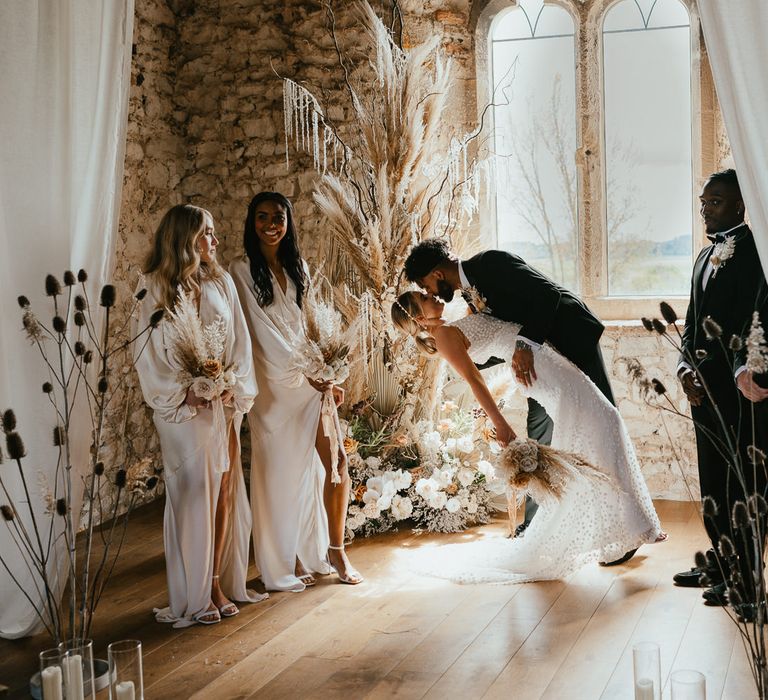 Groom leaning his bride down for a kiss at the wedding ceremony altar surrounded by fresh and dried flower arrangements and drapes. 