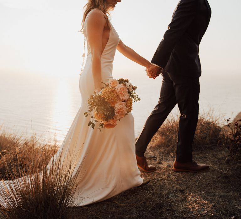 Groom leads bride along the cliffside whilst bride holds peach bouquet