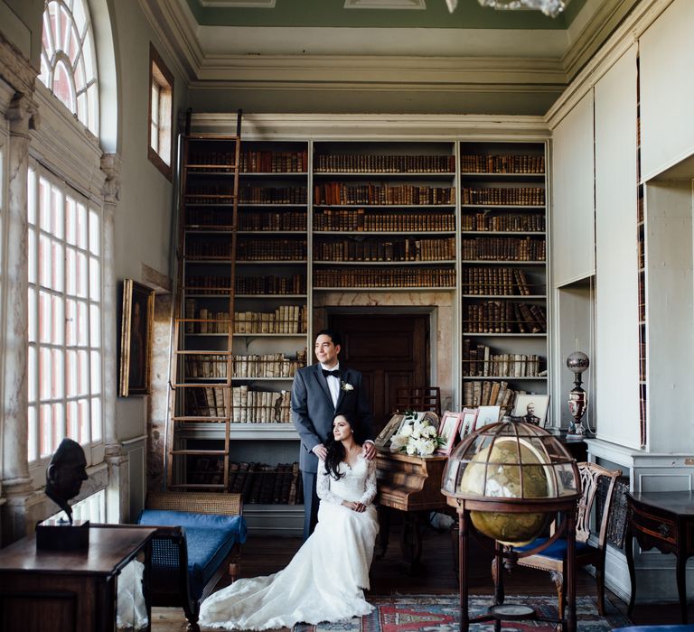 Bride sits down as groom places his hands on her shoulders within regal room filled with books 