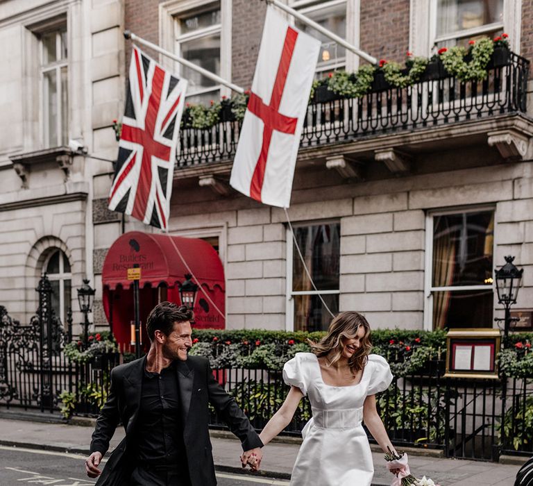 Bride and groom crossing the road in Mayfair, London in an all black suit, and short wedding dress with trailing bow detail 