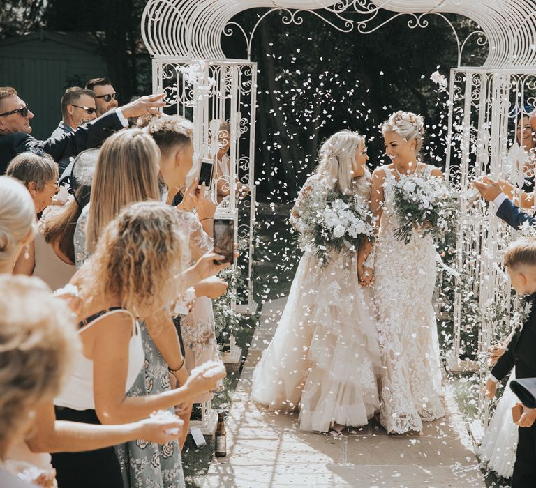 A lesbian couple walk up the aisle after their wedding ceremony through white confetti.