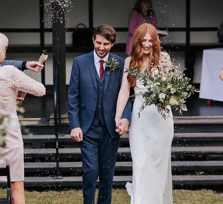 Just married confetti moment with groom in a navy suit holding hands with his bride in a satin slip wedding dress