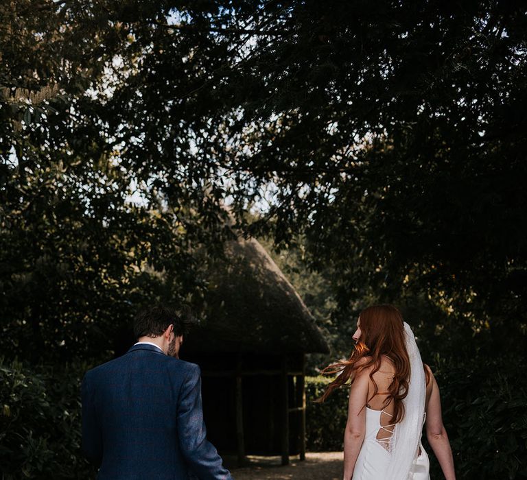 Groom in a navy wool suit holding his brides satin slip wedding dress train and delicate lace cathedral length veil 