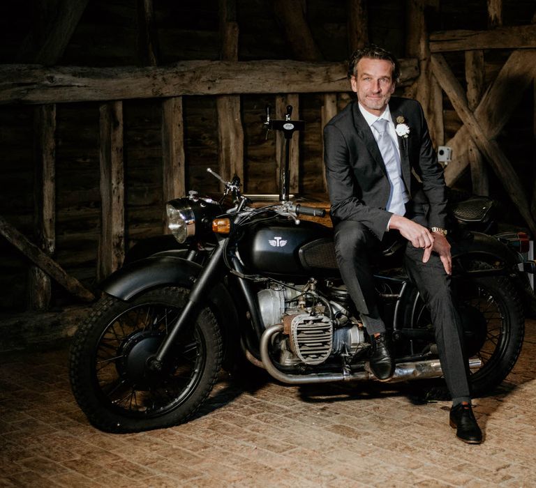 Groom leaning against cocktail bar shaped like a bicycle for rustic luxe wedding 