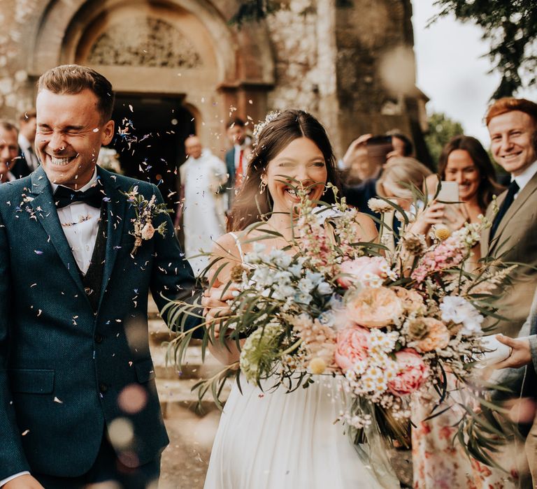 Bride in white cami dress holding multicoloured bridal bouquet walks down the church aisle hand-in-hand with groom in blue suit and bow tie after wedding ceremony