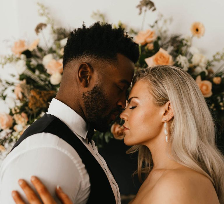 Bride and groom standing nose to nose with each other in front of floral arrangement on a mantlepiece