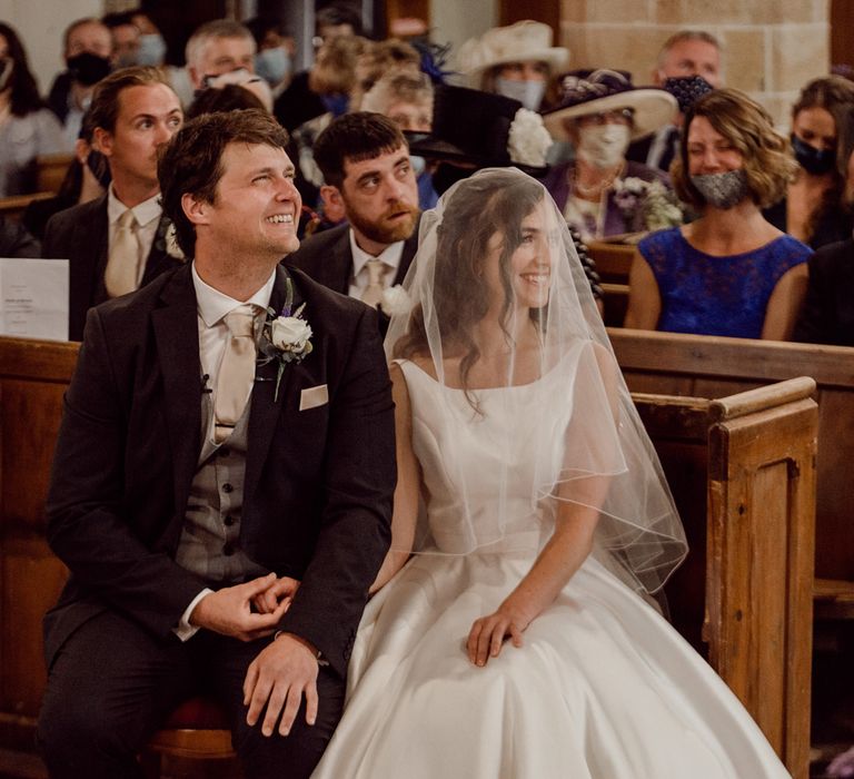 Groom in dark brown three piece suit with grey waistcoat, pale gold tie and pocket square and white rose buttonhole holds hands with bride in white Elbeth Gillis gown and veil as they sit on church pew smiling during wedding ceremony