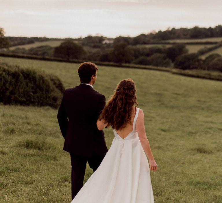 Bride in white low back Elbeth Gillis wedding dress and long curled hair walks arm in arm with groom in dark brown suit through field at home farm wedding