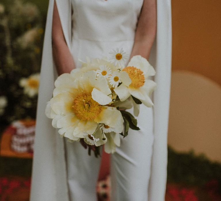 Bride in a jumpsuit and bridal cape holding a white peony and giant daisy wedding bouquet 