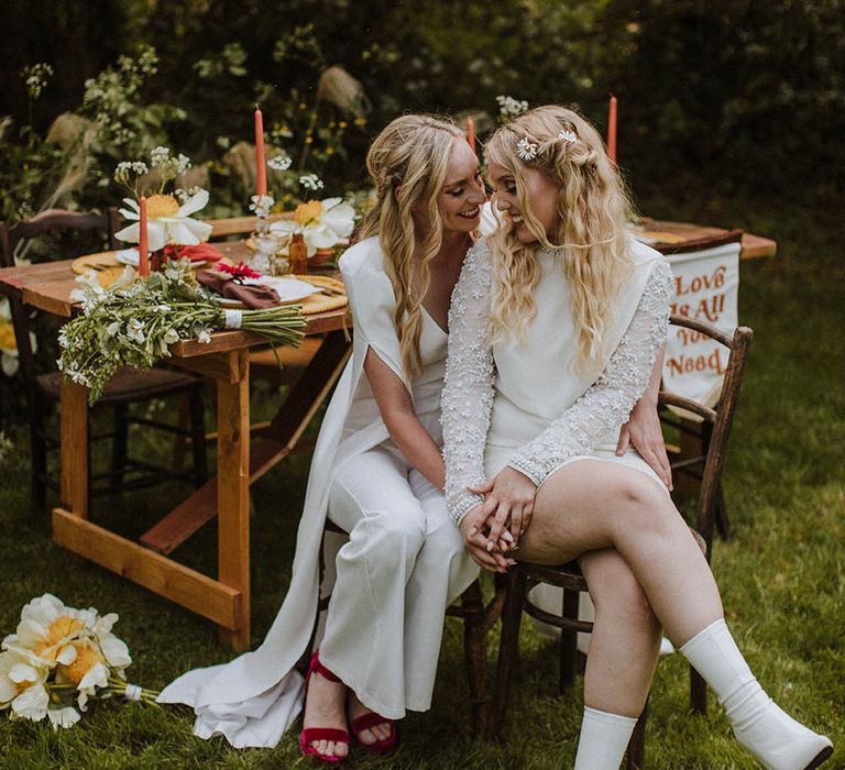 two brides with half up half down wedding hairstyles sitting on a chair at their outdoor sixties wedding 