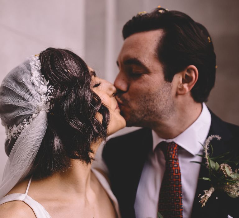 Groom kissing his bride with shoulder length wavy hair wearing a Juliet cap wedding veil 