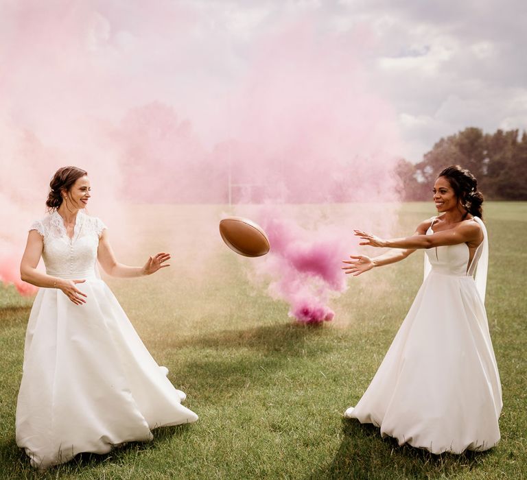 Bride in white cami wedding dress with shoulder veils passes brown rugby ball to bride in white lace top capped sleeved wedding dress on rugby pitch with pink smoke bombs going off in the background after wedding ceremony at The West Mill Derby