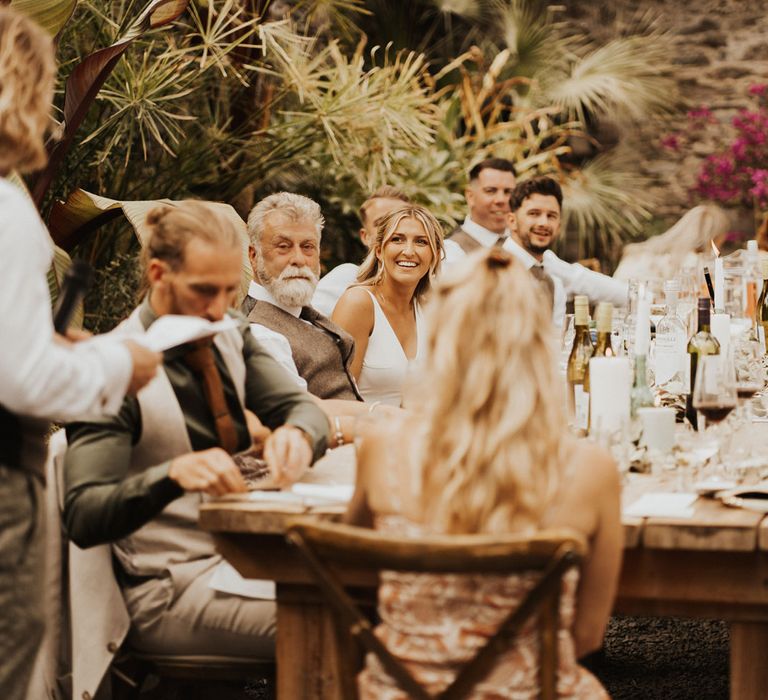 Bride smiles at groom as he makes speech in front of guests at wedding breakfast inside glasshouse at Anran Devon with wooden tables, candles and palm leaves