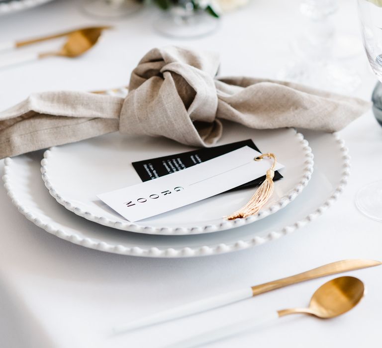 A neutral toned napkin is tied elegantly and rests on a white top plate. A guest name tag rests next to it. the tag is white and has a gold tassel. the cuttlery is gold and white.