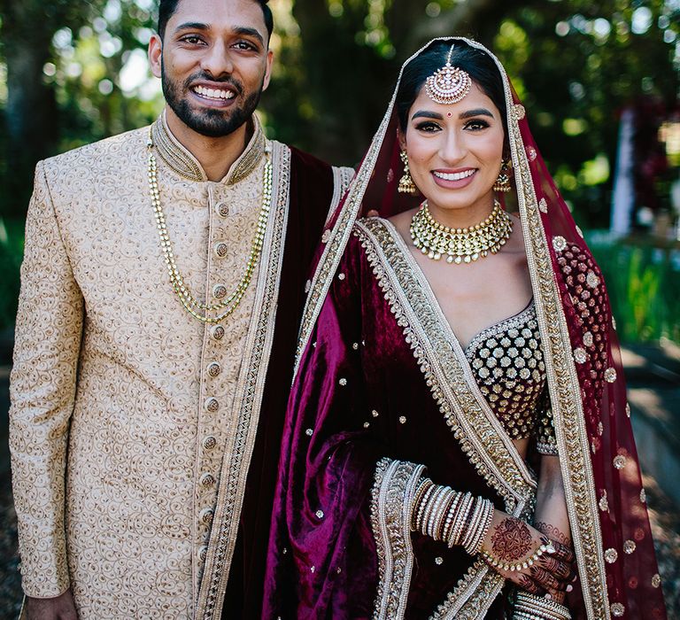 An Indian bride stands with her husband wearing traditional dress in dark red. He wears a gold suit. 