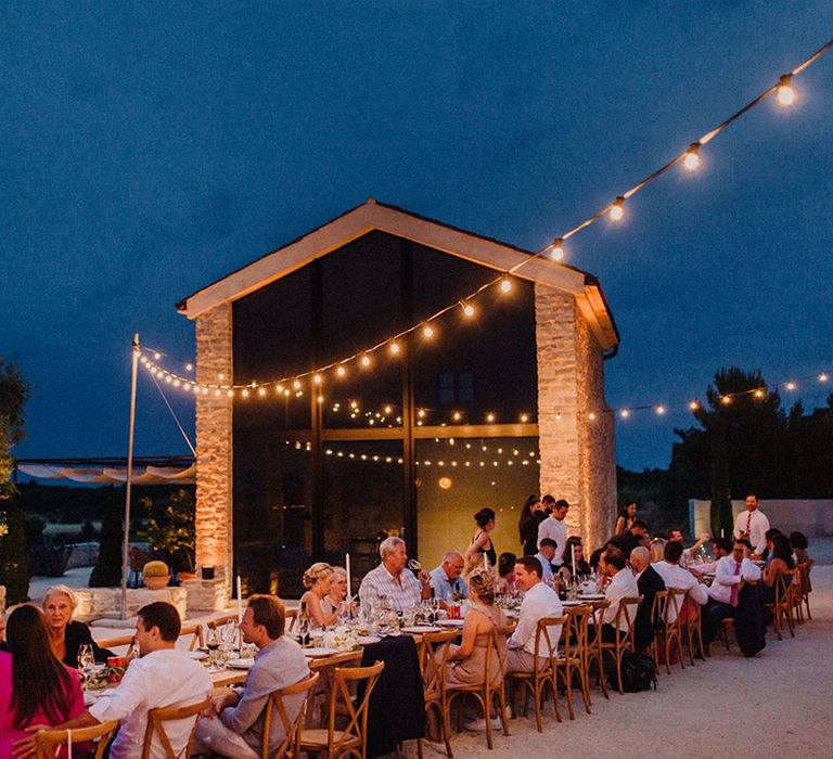 Wedding party sit outdoors as fairy lights hang above rustic table setting