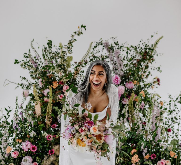 Bride in satin Halfpenny London wedding dress and tasselled bridal cape holding multicoloured bridal bouquet stands in front of multicoloured floral installation for wedding at Loft Studios London