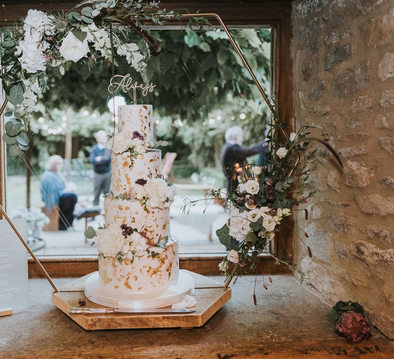 White four tier wedding cake with gold details decorated with white roses, green leaves and cake topper reading 'Always' on gold hexagonal cake stand at Tythe Barn wedding with barn wedding flowers