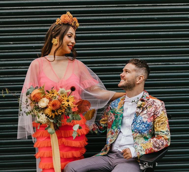 Bride in a coral wedding dress and cape veil holding a yellow and orange wedding bouquet stood next to her disabled groom in a wheelchair wearing a brightly coloured jacket 