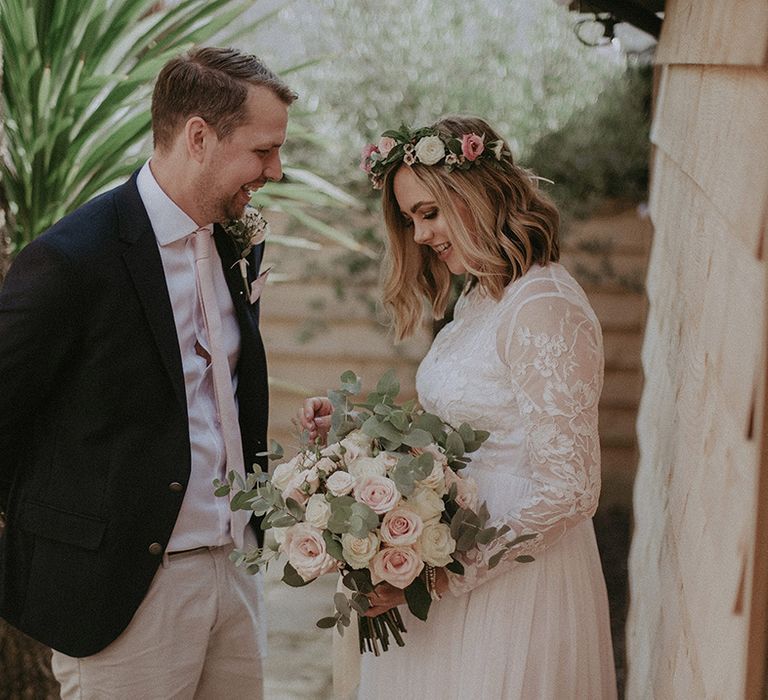 Bride in a long sleeve lace and tulle wedding dress with white and pink flower crown and bouquet laughing with her husband in a navy blazer and beige shorts 