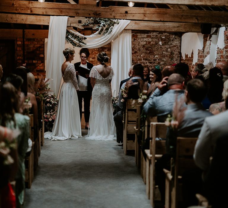 Two brides standing at the altar of their Woolas Barn wedding with drapes and fairy lights decorating the altar 