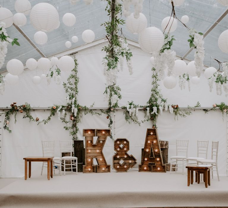 Interior of marquee wedding reception with stage, large wooden light-up letters, chairs, garlands hanging from the ceiling and white paper lanterns for garden wedding in Bedfordshire