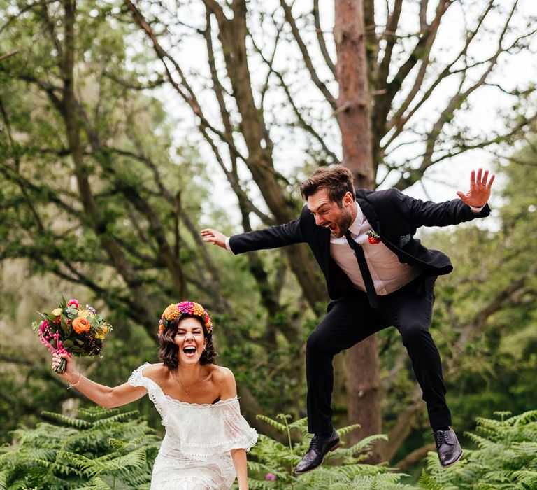 Bride in lace Grace Loves Lace wedding dress, flower crown and holding colourful bridal bouquet jumps in the woods with groom in black suit at The Bridal Barn venue