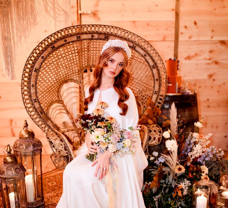 Bride in white cape and wedding dress with white netted headpiece holding colourful bridal bouquet t sits on large rattan chair surrounded by boho decor