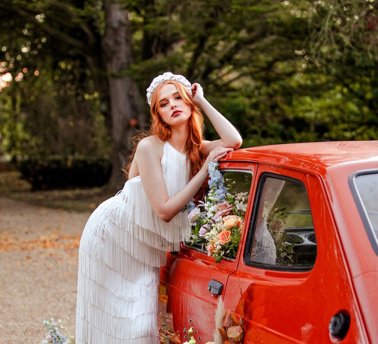 Bride in long fringed wedding dress and white headband leans on red car filled with colourful florals after marquee tent wedding