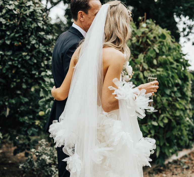 Bride in Halfpenny London wedding dress and Jimmy Choo wedding shoes walks with groom whilst holding Halfpenny London ruffle veil and champagne flute