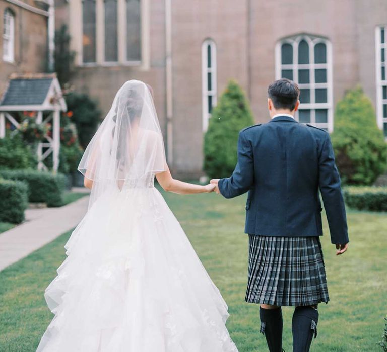 Groom in a blue tartan kilt and blazer holding hands with his bride in a princess wedding dress with layered tulle skirt at Scottish castle wedding 