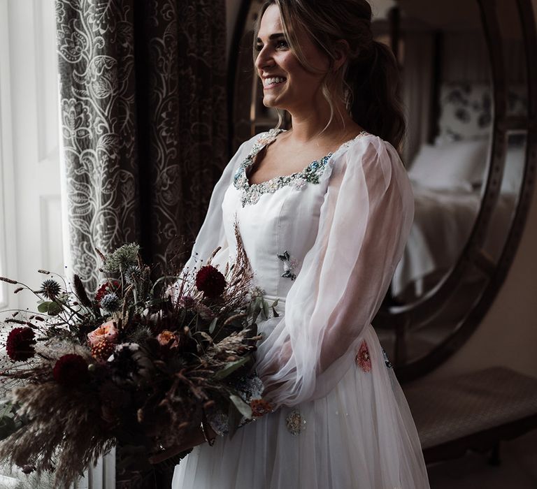 Bride wearing long mesh sleeve wedding dress and dried autumnal wedding bouquet stands by window before wedding