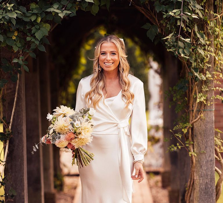 Bride in white satin dress with train, vail and long sleeve satin wrap top stands under pergola holding white, yellow and pink wedding bouquet 