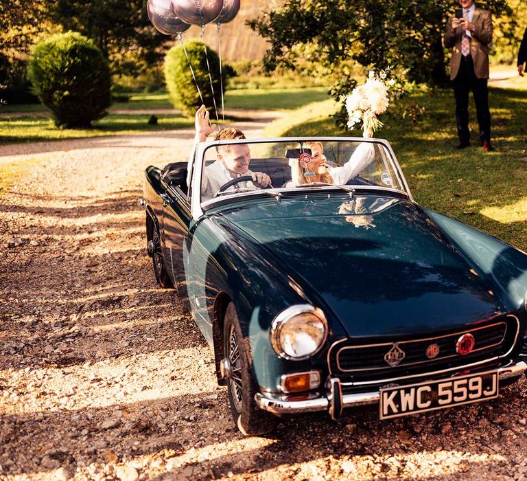 Bride holding up bouquet and groom in grey suit waving his hand sit in vintage teal convertible with balloons on the back as they drive out of churchyard