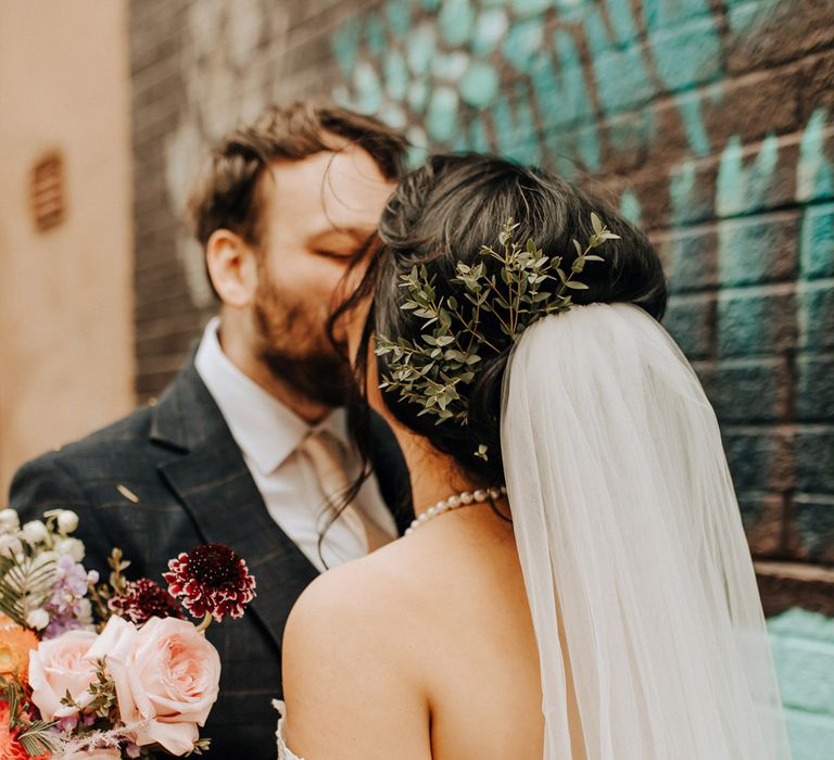 Bride in lace drop strap wedding dress with foliage hair decor and veil kisses groom by street art in Liverpool after wedding ceremony