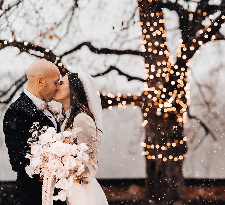 Groom in a black suit kissing his bride in a long sleeve wedding dress as she holds an all-white wedding bouquet 