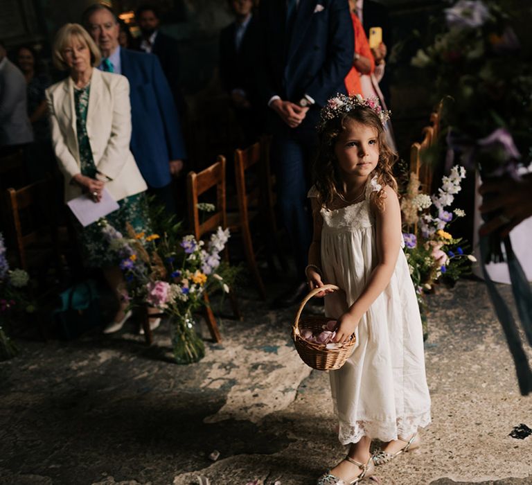 Flower girl in white dress and flower crown throws pink rose petals from a wicker basket 