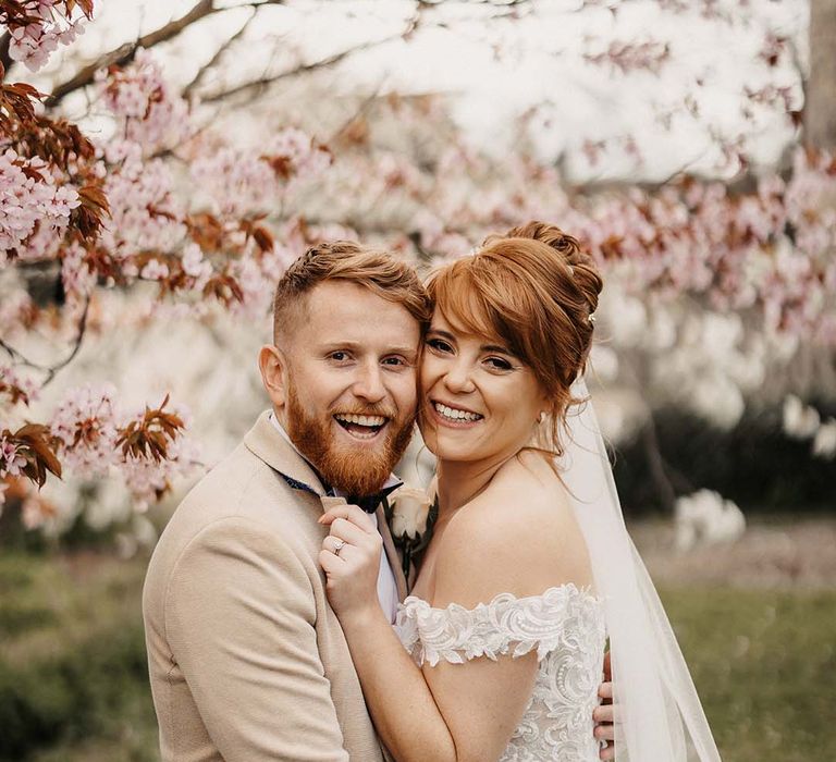 The happy newlyweds stand under pink blossom trees after their wedding ceremony in rustic barn wedding venue