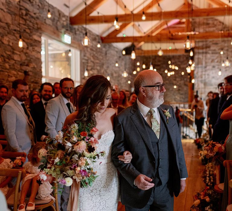 Father of the bride walks bride down the aisle in grey suit and paisley green tie with bride holding wildflower wedding bouquet